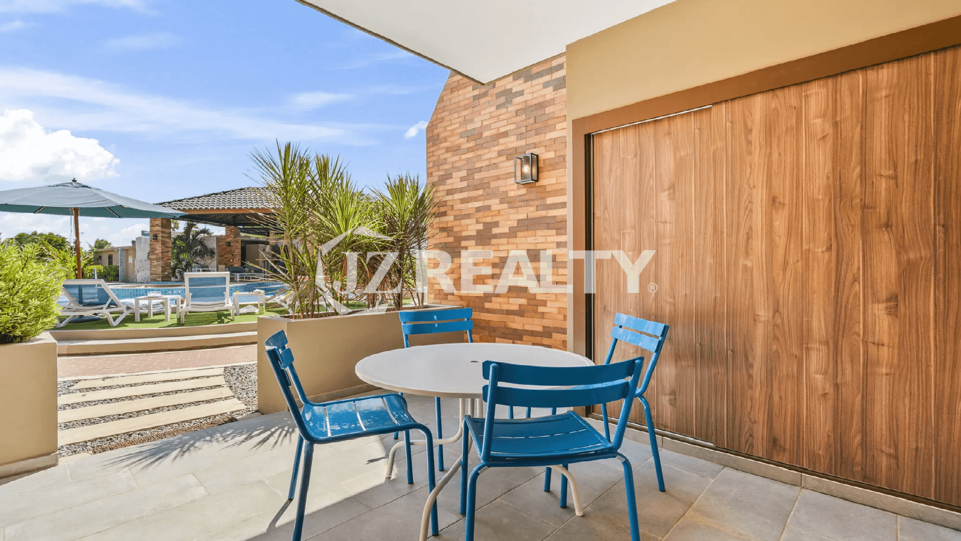 Covered ground floor patio with an outdoor dining set (blue chairs) and direct access/view of the Porta al Sole community pool area in Noord, Aruba.