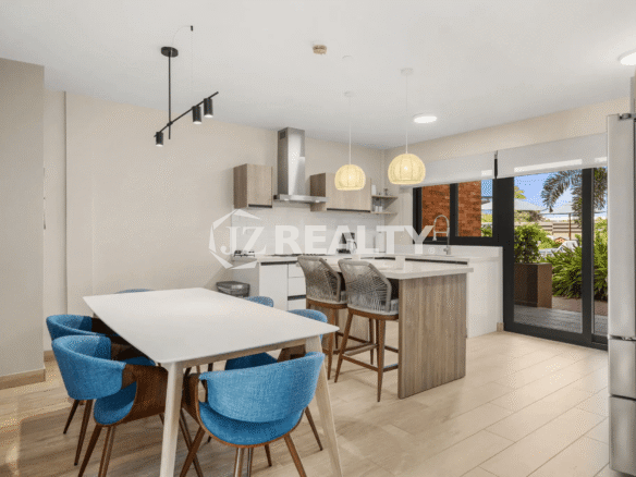 Dining area in Porta al Sole Unit 12 with a large white table and blue chairs, adjacent to the modern kitchen with a center island and view to the outdoor patio.