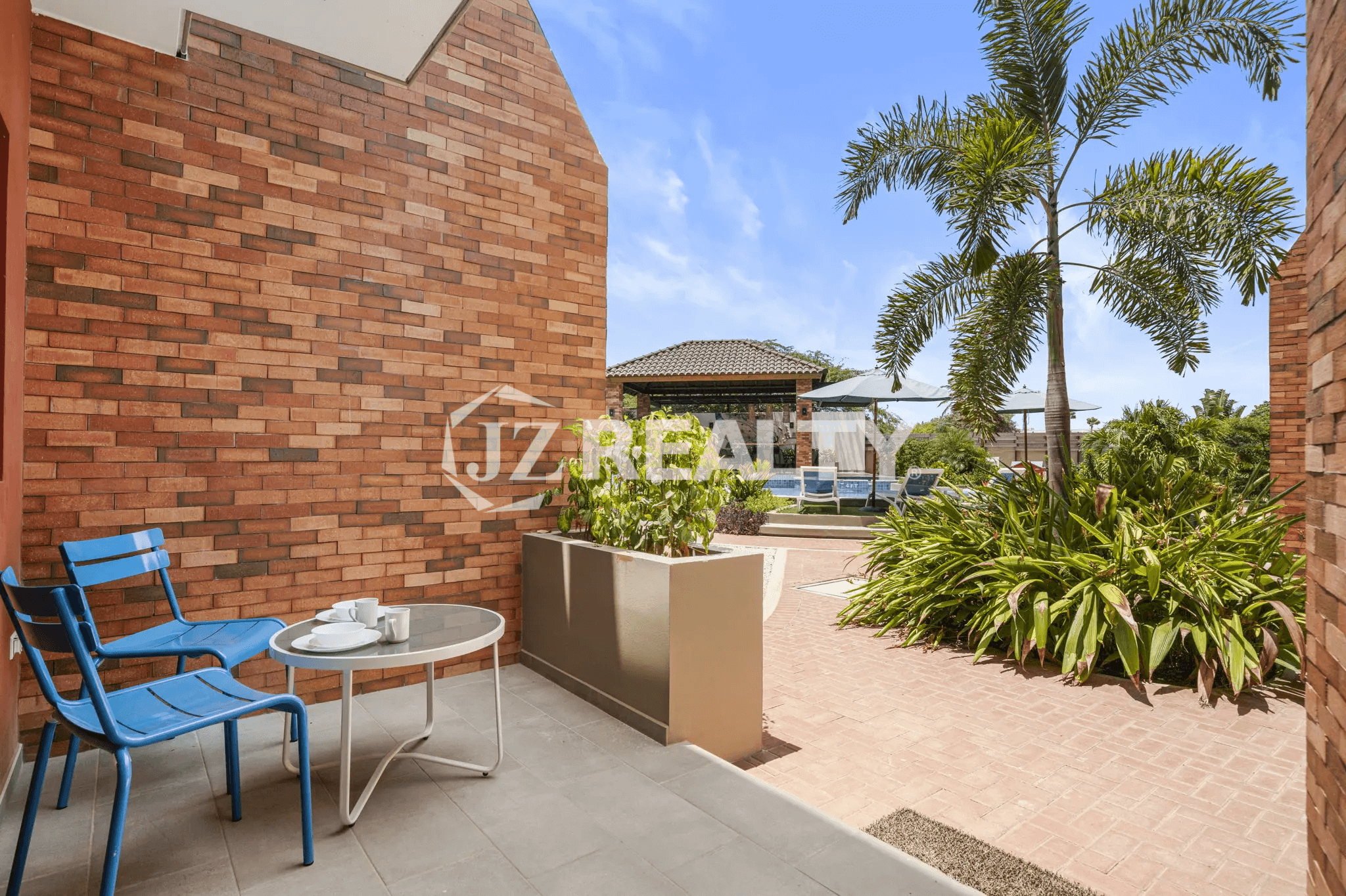 Private ground-floor patio area of Porta al Sole Unit 12 with a brick wall backdrop, outdoor table and blue chairs, and tropical planter box.
