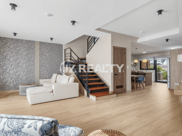 Open-plan living area in Porta al Sole Unit 12 with light wood flooring, a white sectional sofa, decorative wall paneling, and an open modern staircase leading to the second floor.