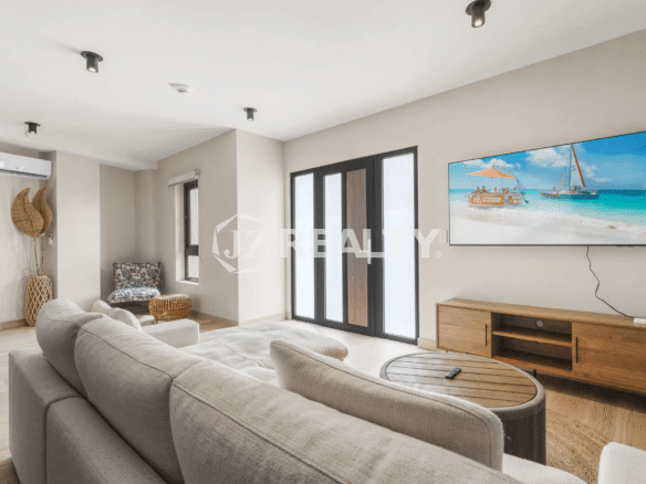 Cozy and modern living room in Porta al Sole Unit 12 with a large L-shaped light gray sofa, wall-mounted TV displaying a beach scene, wood media console, and black-framed patio doors.