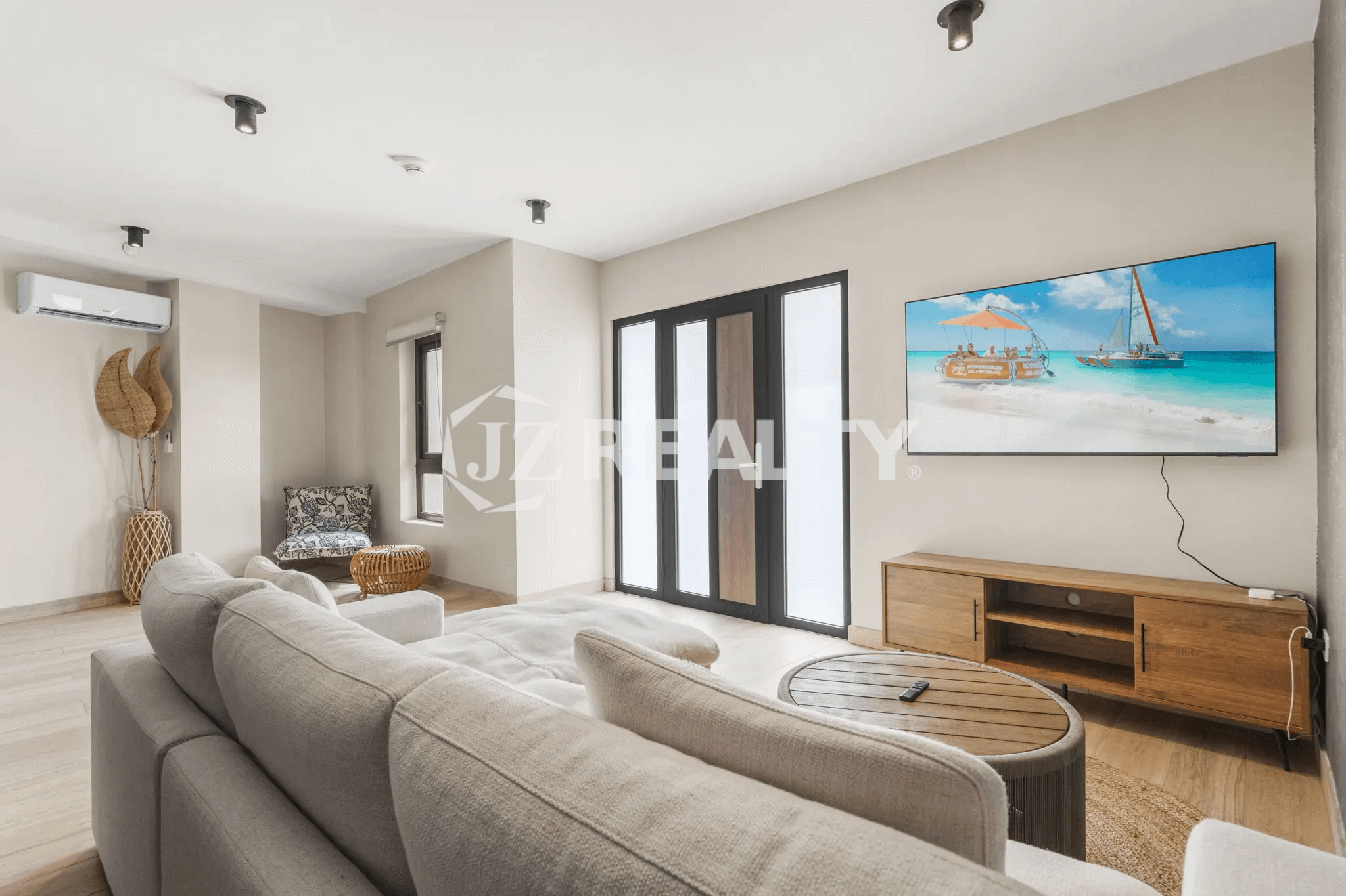 Cozy and modern living room in Porta al Sole Unit 12 with a large L-shaped light gray sofa, wall-mounted TV displaying a beach scene, wood media console, and black-framed patio doors.