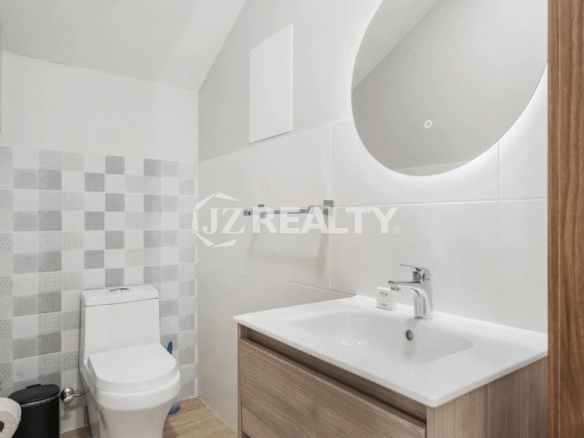 Contemporary bathroom in Porta al Sole Unit 12 featuring a floating wooden vanity, white sink, backlit circular mirror, and patterned accent tile wall behind the toilet.