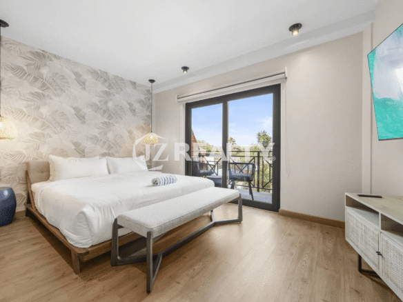 Bright primary bedroom in Porta al Sole Unit 12 with a king-sized bed, tropical leaf wallpaper accent wall, wood flooring, black-framed sliding glass door to a balcony, and a wall-mounted TV.