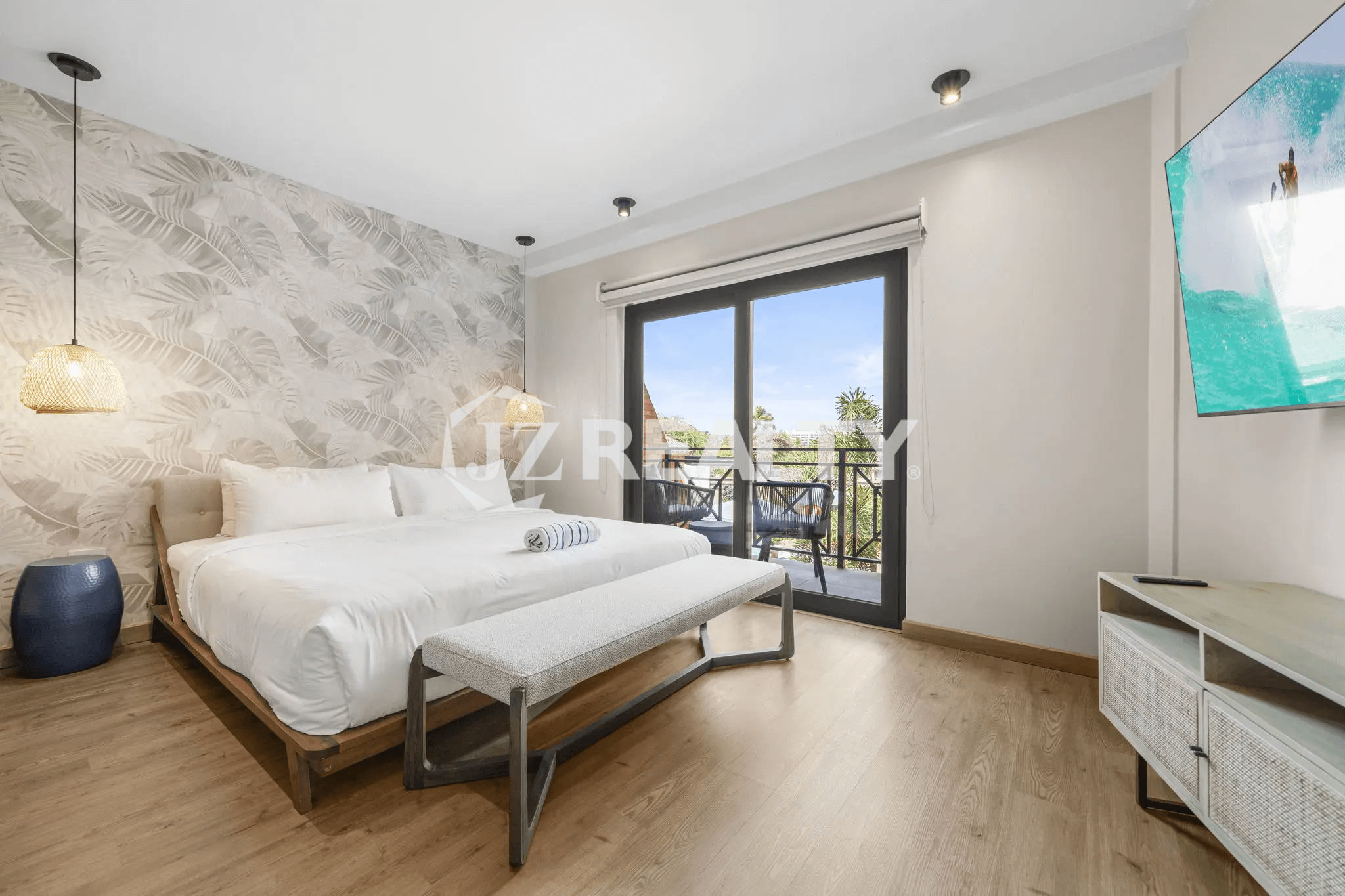 Bright primary bedroom in Porta al Sole Unit 12 with a king-sized bed, tropical leaf wallpaper accent wall, wood flooring, black-framed sliding glass door to a balcony, and a wall-mounted TV.