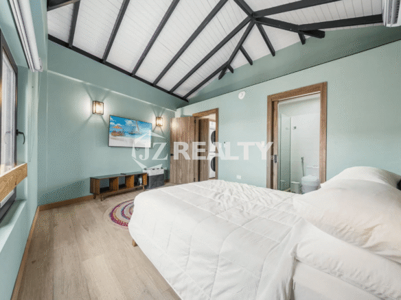 Bedroom in Porta al Sole Unit 12 with light green walls, a vaulted ceiling with exposed beams, a queen-sized bed, a wall-mounted TV, and an entrance to an ensuite bathroom.