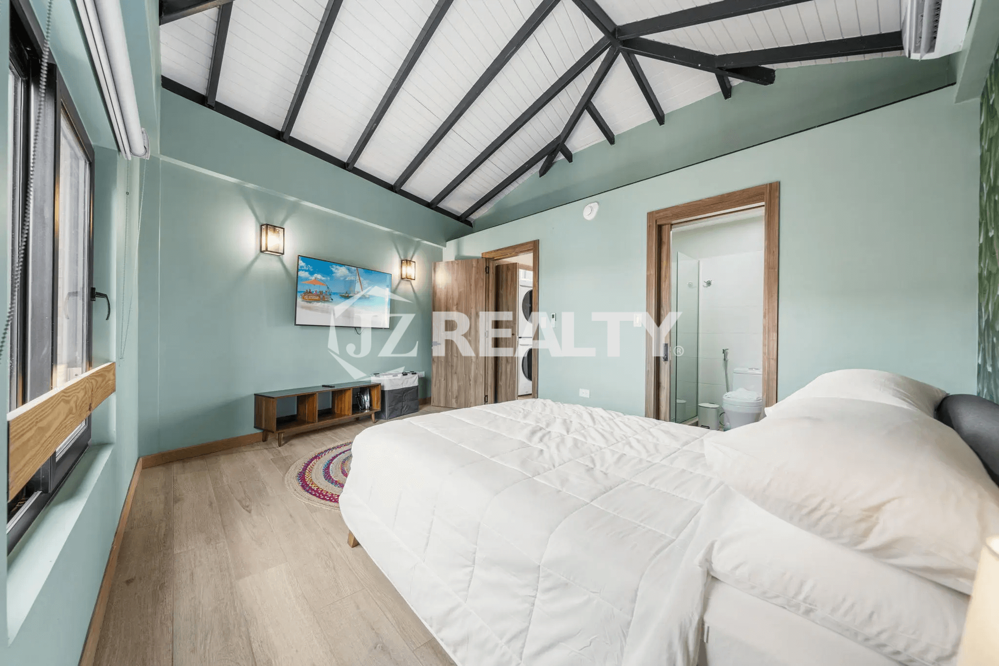 Bedroom in Porta al Sole Unit 12 with light green walls, a vaulted ceiling with exposed beams, a queen-sized bed, a wall-mounted TV, and an entrance to an ensuite bathroom.