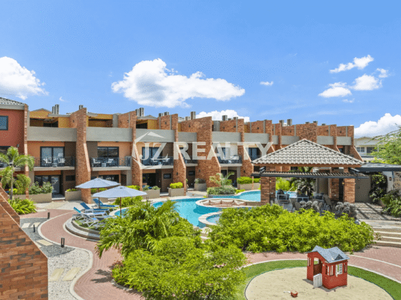 Wide view of the property's central courtyard, showing the multi-story townhouses with balconies, the main pool area, surrounding landscaping, and a small children's playground/sandpit.