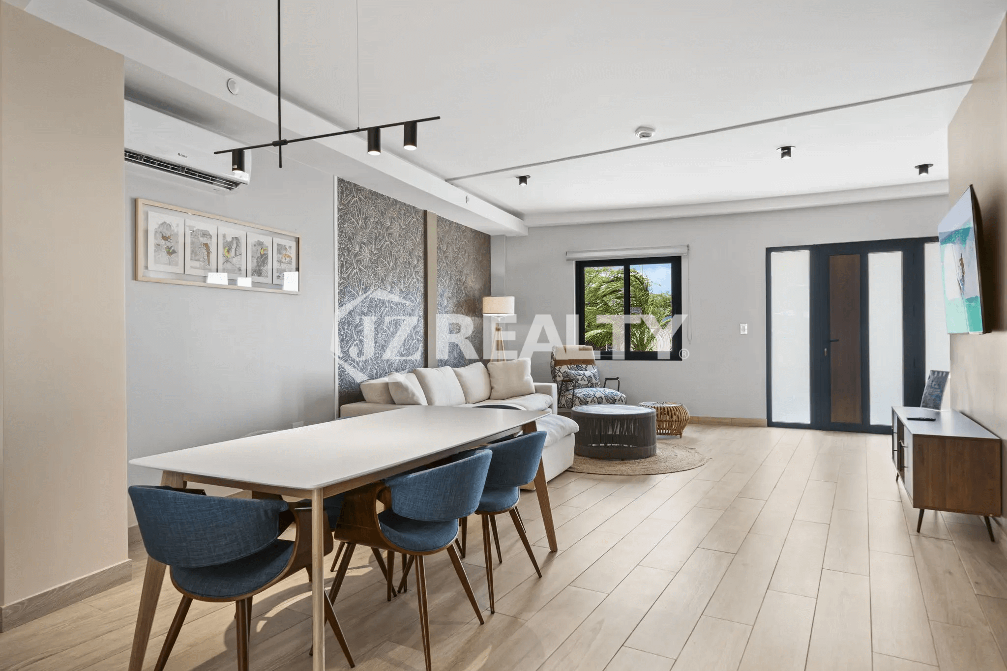 Open concept dining and living area in Porta al Sole Unit 13 with long white dining table, blue chairs, white sofa, and light wood floors.