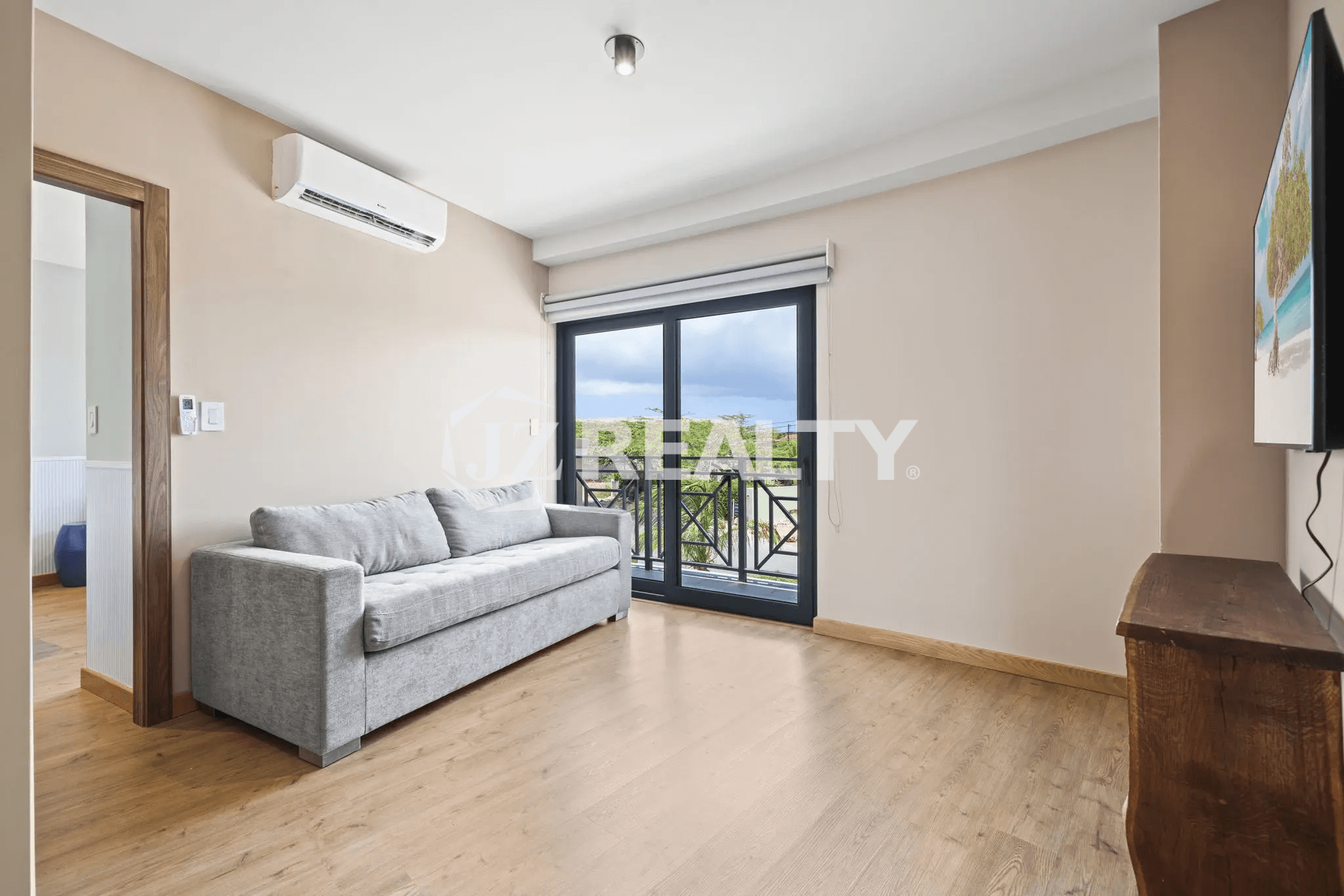 Interior view of a living room in Porta al Sole Unit 13 featuring a grey sofa, hardwood floor, AC unit, and glass door leading to a balcony.