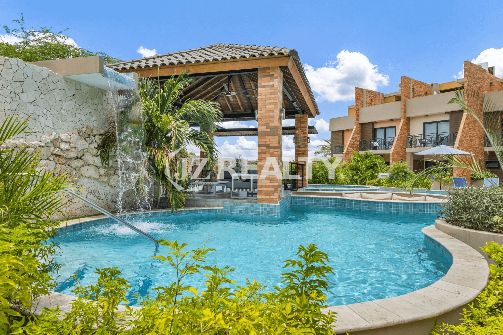 View of the swimming pool and attached jacuzzi/hot tub, featuring a waterfall flowing from a stone-textured wall and a covered wooden palapa with outdoor seating in the background.