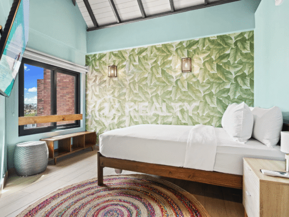 Bright bedroom with a tropical leaf accent wall, exposed ceiling beams, and a round woven rug in Porta al Sole Unit 14.