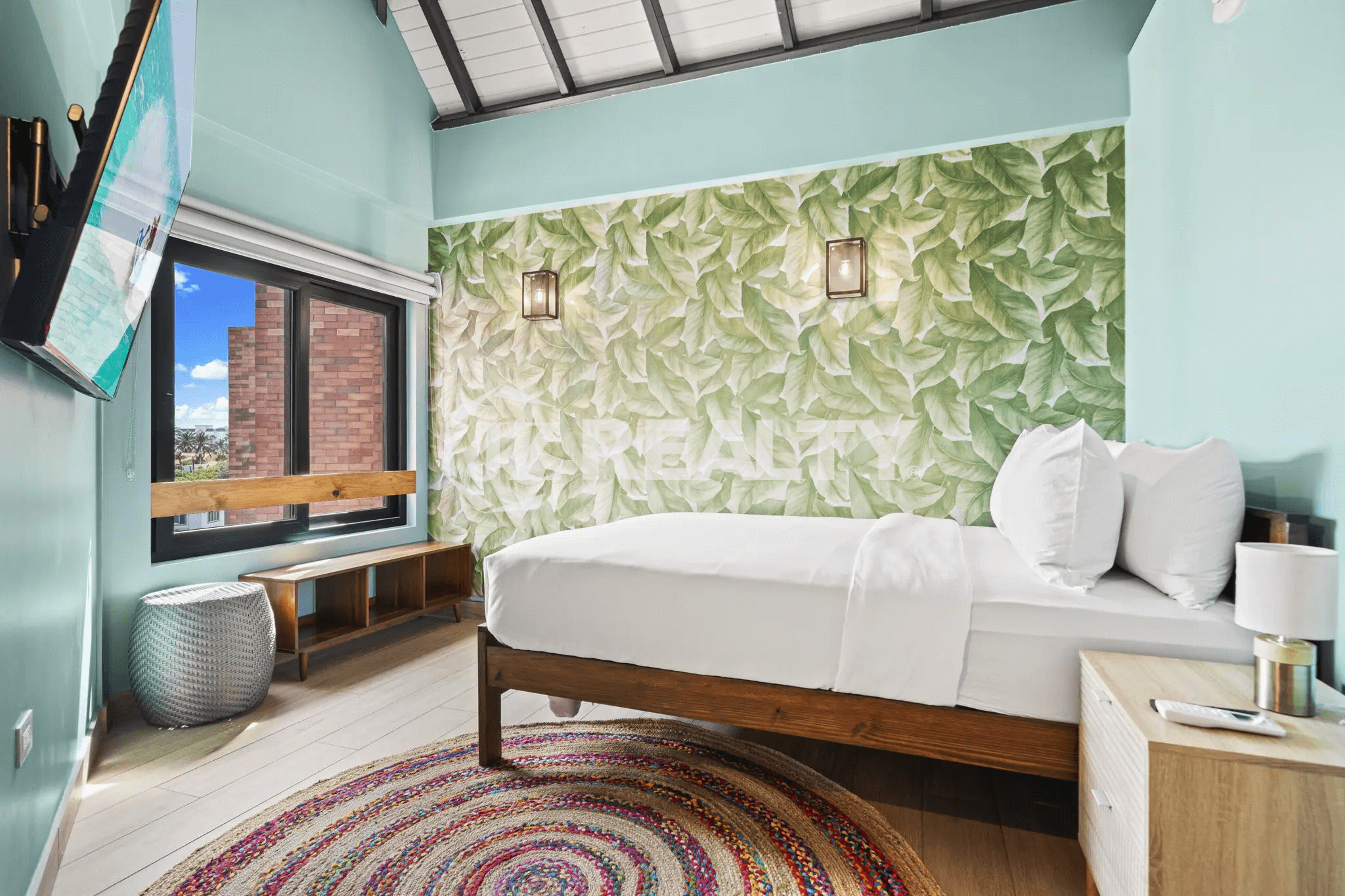 Bright bedroom with a tropical leaf accent wall, exposed ceiling beams, and a round woven rug in Porta al Sole Unit 14.