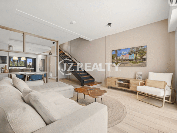 Wide-angle view of the bright, modern living room with a large TV, light wood media console, comfortable seating, and the sleek metal and wood open staircase in Porta al Sole Unit 15.