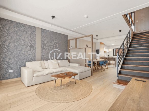 View of the contemporary open-plan living room with a large light-colored sectional sofa, dark patterned accent wall, and wood-look flooring, leading toward the dining area and stairwell in Porta al Sole Unit 15.