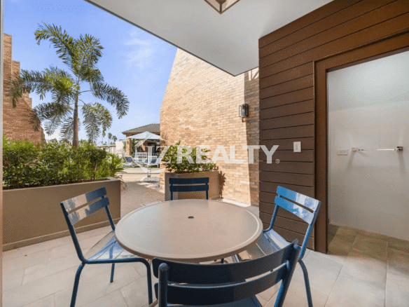 Outdoor dining patio with a small table and blue chairs, surrounded by brick walls, tropical plants, and a view of the common pool area in Porta al Sole Unit 15.