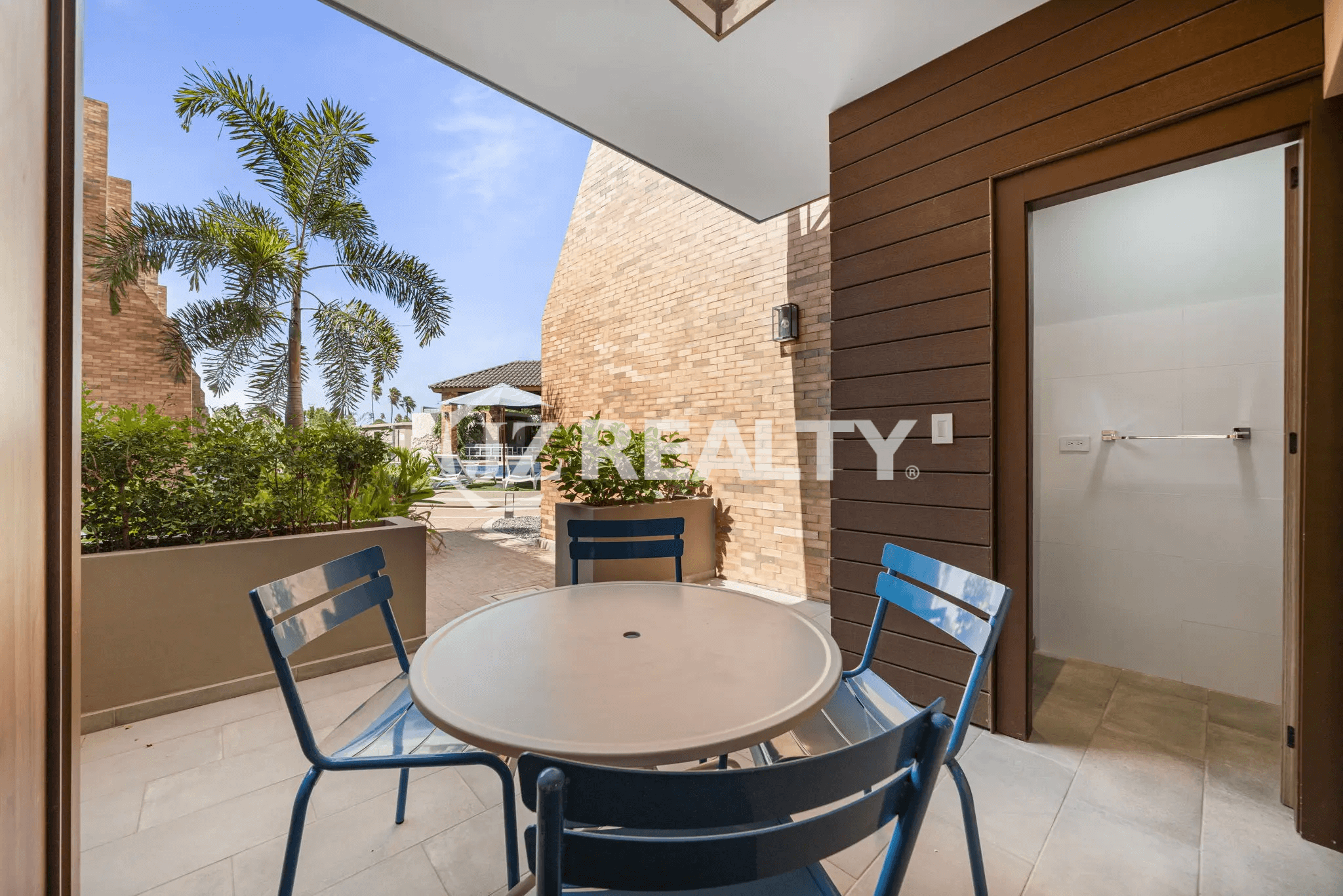 Outdoor dining patio with a small table and blue chairs, surrounded by brick walls, tropical plants, and a view of the common pool area in Porta al Sole Unit 15.