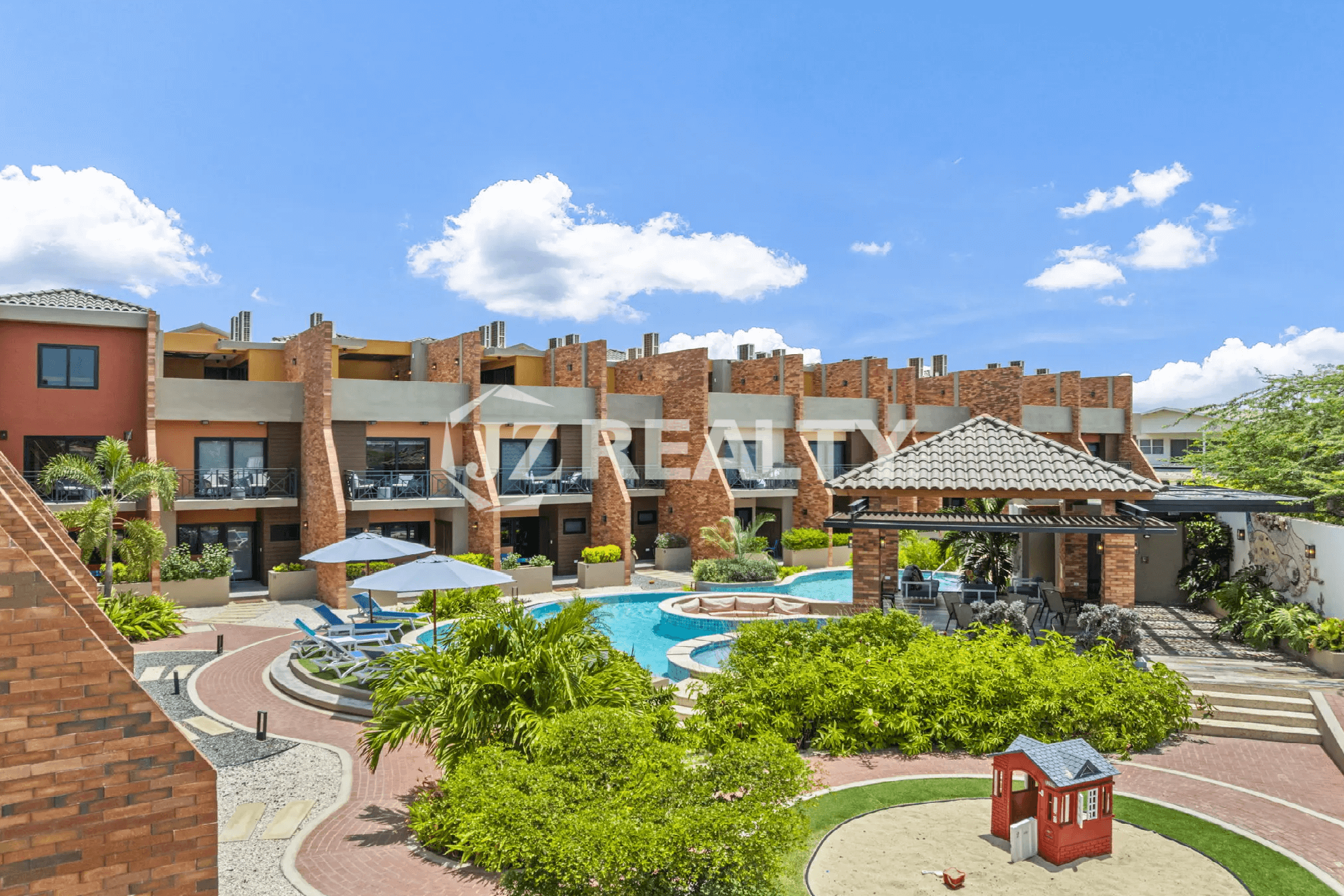 Elevated view of the Porta al Sole community, showcasing the central swimming pool, landscaped grounds, lounge chairs, a small children's play area, and the surrounding townhouse buildings.