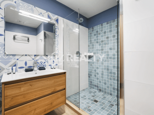Contemporary bathroom with a light wood vanity, white sink, large mirror, and a glass-enclosed shower featuring blue and white mosaic tiles.