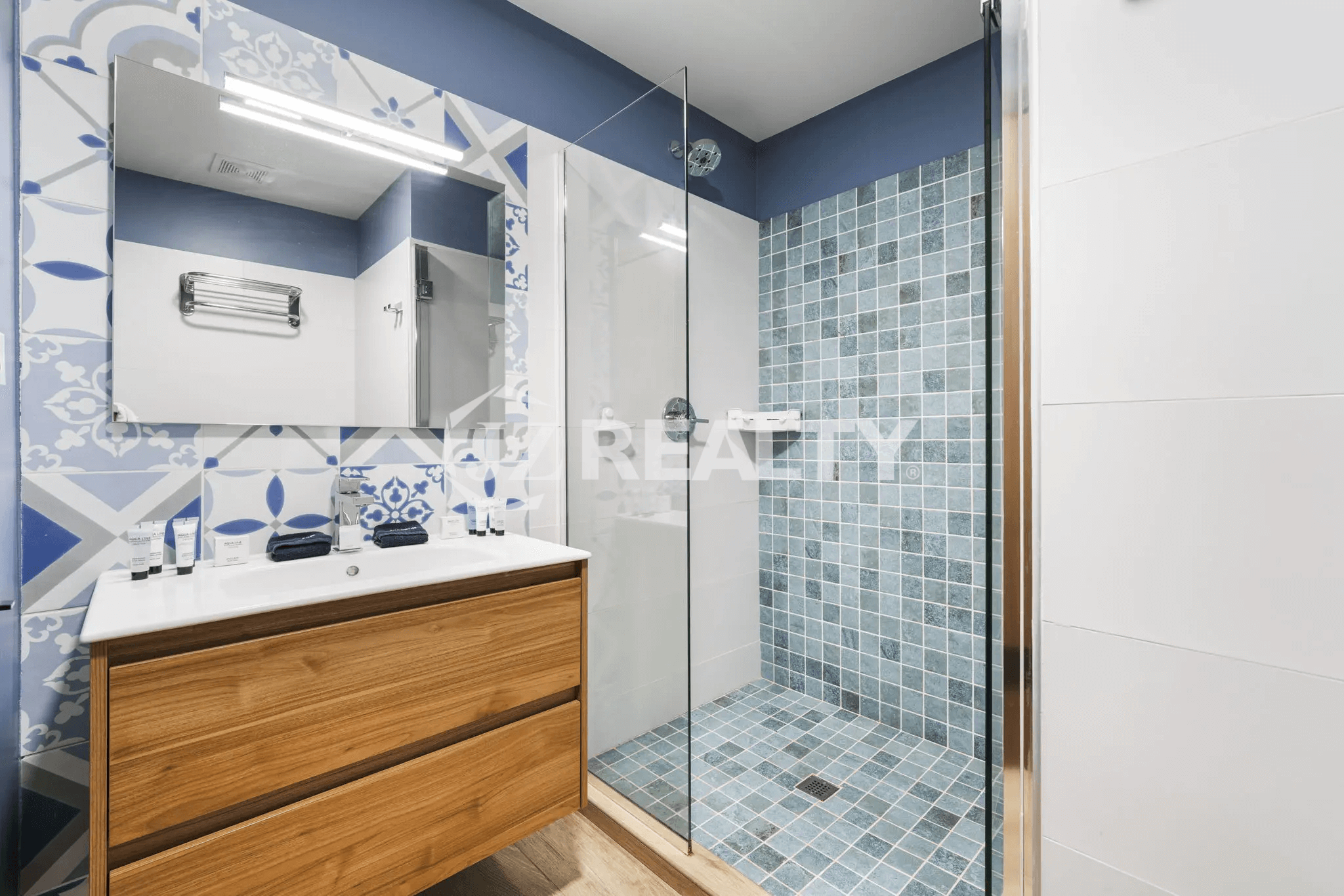 Contemporary bathroom with a light wood vanity, white sink, large mirror, and a glass-enclosed shower featuring blue and white mosaic tiles.