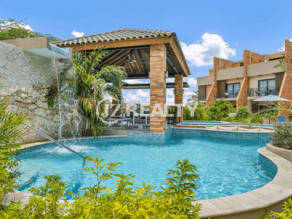 Close-up view of the community pool with built-in seating, a stone waterfall, and a nearby covered cabana with brick pillars and lounge seating.