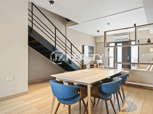 Dining area with a long wooden table and modern blue chairs, adjacent to a contemporary black metal staircase; part of the living area is visible through a decorative wood-framed divider.