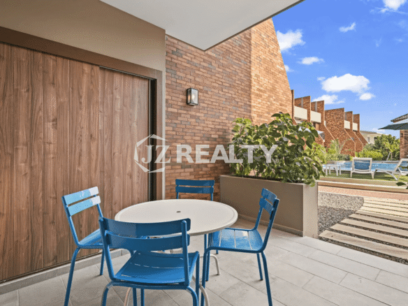 Private ground-floor patio area with a white round table and four bright blue chairs, brick accent wall, and wood-look flooring, looking out towards the communal pool area.