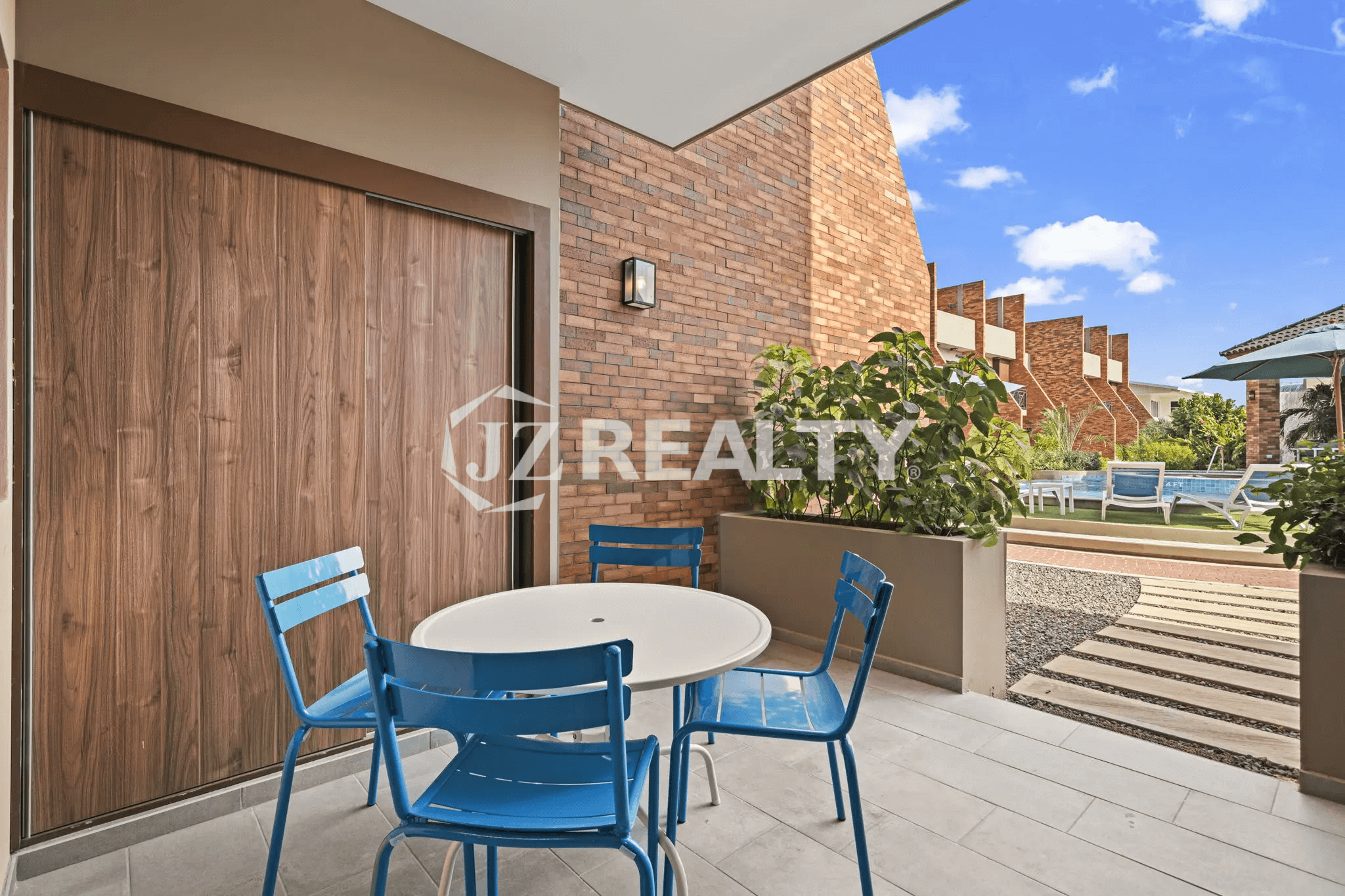 Private ground-floor patio area with a white round table and four bright blue chairs, brick accent wall, and wood-look flooring, looking out towards the communal pool area.