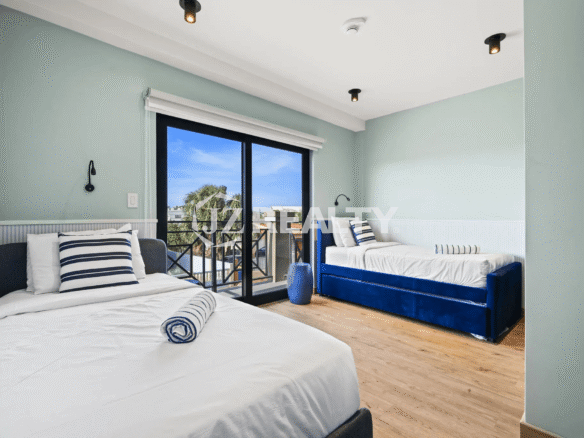 Guest bedroom with light blue walls, two beds with navy and white striped pillows (one twin, one daybed-style), wood-look flooring, and a sliding glass door leading to a small balcony.