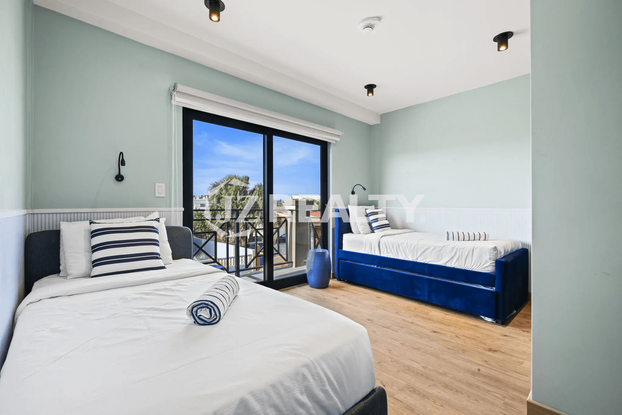 Guest bedroom with light blue walls, two beds with navy and white striped pillows (one twin, one daybed-style), wood-look flooring, and a sliding glass door leading to a small balcony.