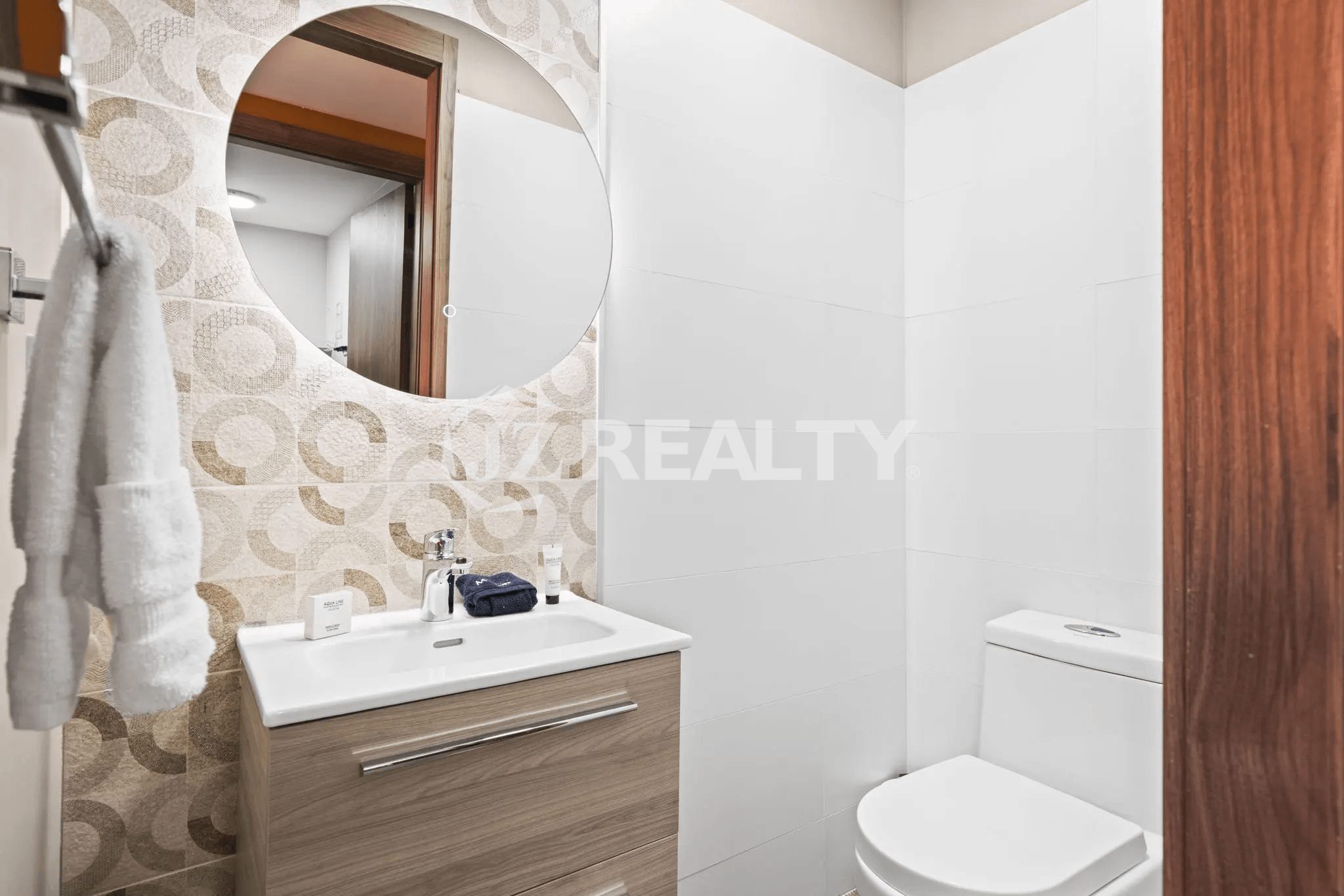 Small white bathroom with a circular mirror, white sink on a wooden vanity, a toilet, and patterned beige and white accent wall tiles.