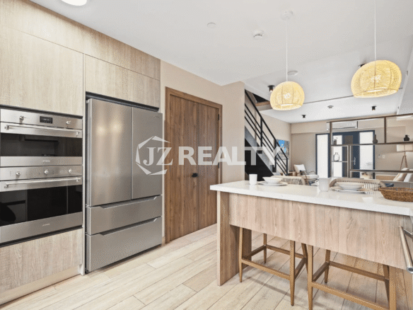 Modern kitchen cabinetry featuring light wood veneer, stainless steel double oven, and a large stainless steel refrigerator, with a view toward the main living area.