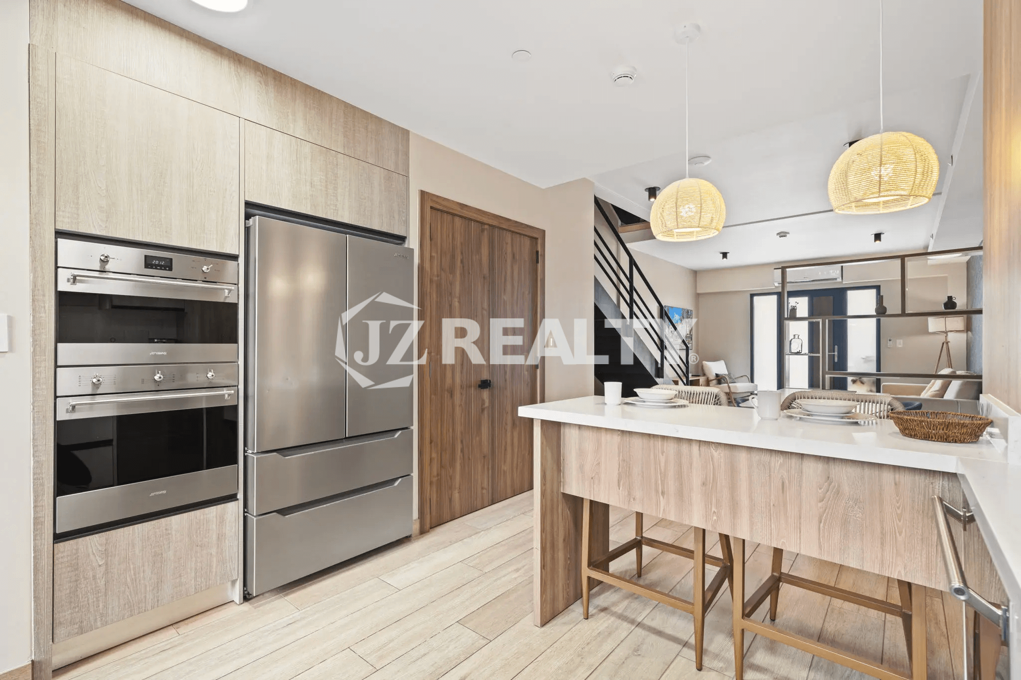 Modern kitchen cabinetry featuring light wood veneer, stainless steel double oven, and a large stainless steel refrigerator, with a view toward the main living area.
