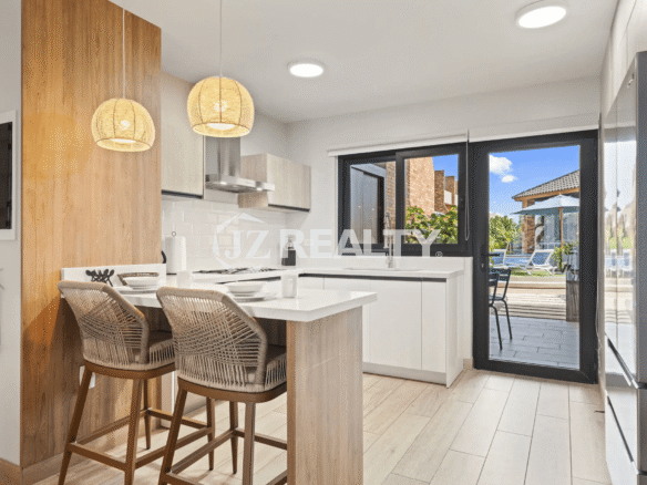Contemporary kitchen with white lower cabinets, light wood accents, a small breakfast bar/island with rattan stools, and a glass door providing direct access to the outdoor patio and pool area.