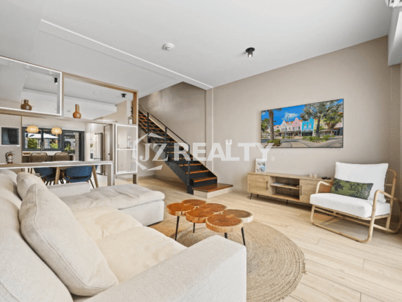 Modern living room with a beige sectional sofa, rustic wood slice coffee table, white accent chair, and a wall-mounted TV; the staircase and dining area are visible beyond a decorative divider.