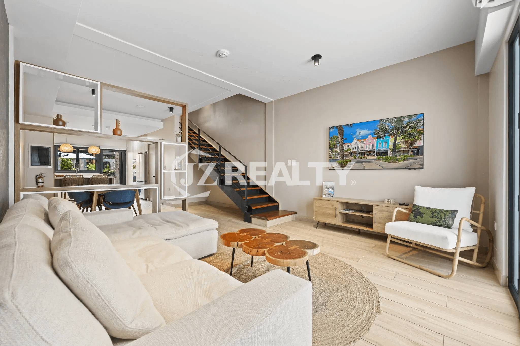 Modern living room with a beige sectional sofa, rustic wood slice coffee table, white accent chair, and a wall-mounted TV; the staircase and dining area are visible beyond a decorative divider.