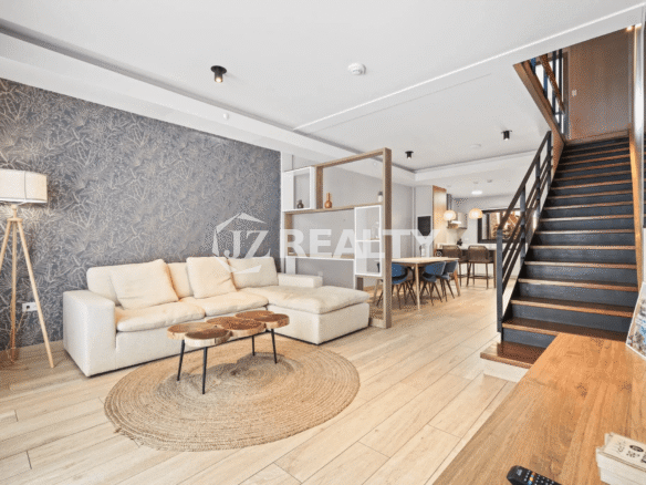 Spacious open-concept living room with a large off-white sectional sofa, round wooden coffee table, jute rug, textured accent wall, and wood flooring leading to the dining area and staircase.