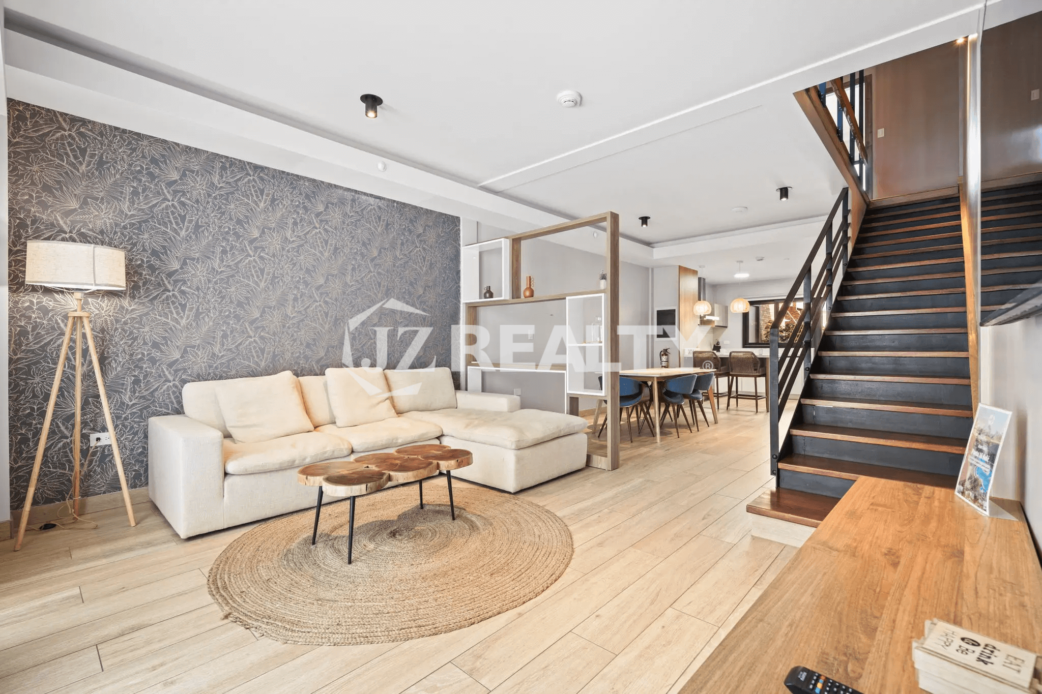 Spacious open-concept living room with a large off-white sectional sofa, round wooden coffee table, jute rug, textured accent wall, and wood flooring leading to the dining area and staircase.