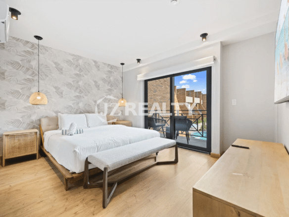 Primary bedroom featuring a large white bed, tropical-patterned accent wall, woven pendant lights, wood flooring, and sliding glass doors leading to a private balcony overlooking the pool.