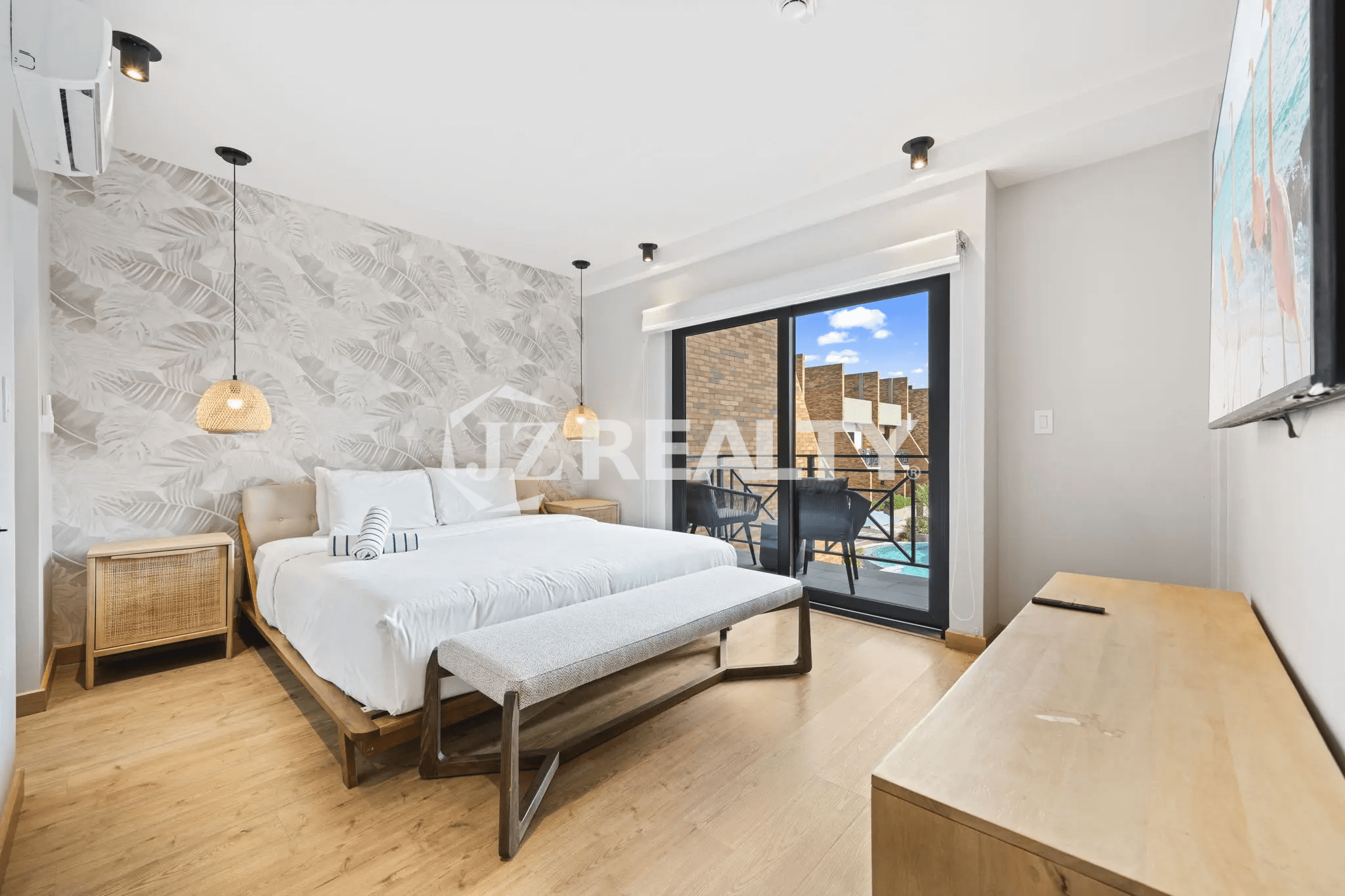 Primary bedroom featuring a large white bed, tropical-patterned accent wall, woven pendant lights, wood flooring, and sliding glass doors leading to a private balcony overlooking the pool.