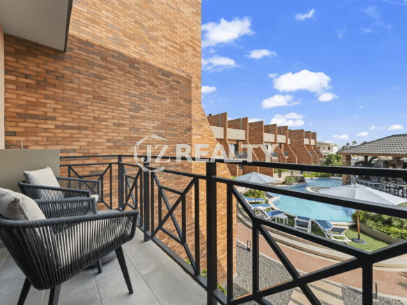 Private outdoor balcony with black metal railings, two modern rope chairs, and a view of the shared pool area and surrounding brick buildings.