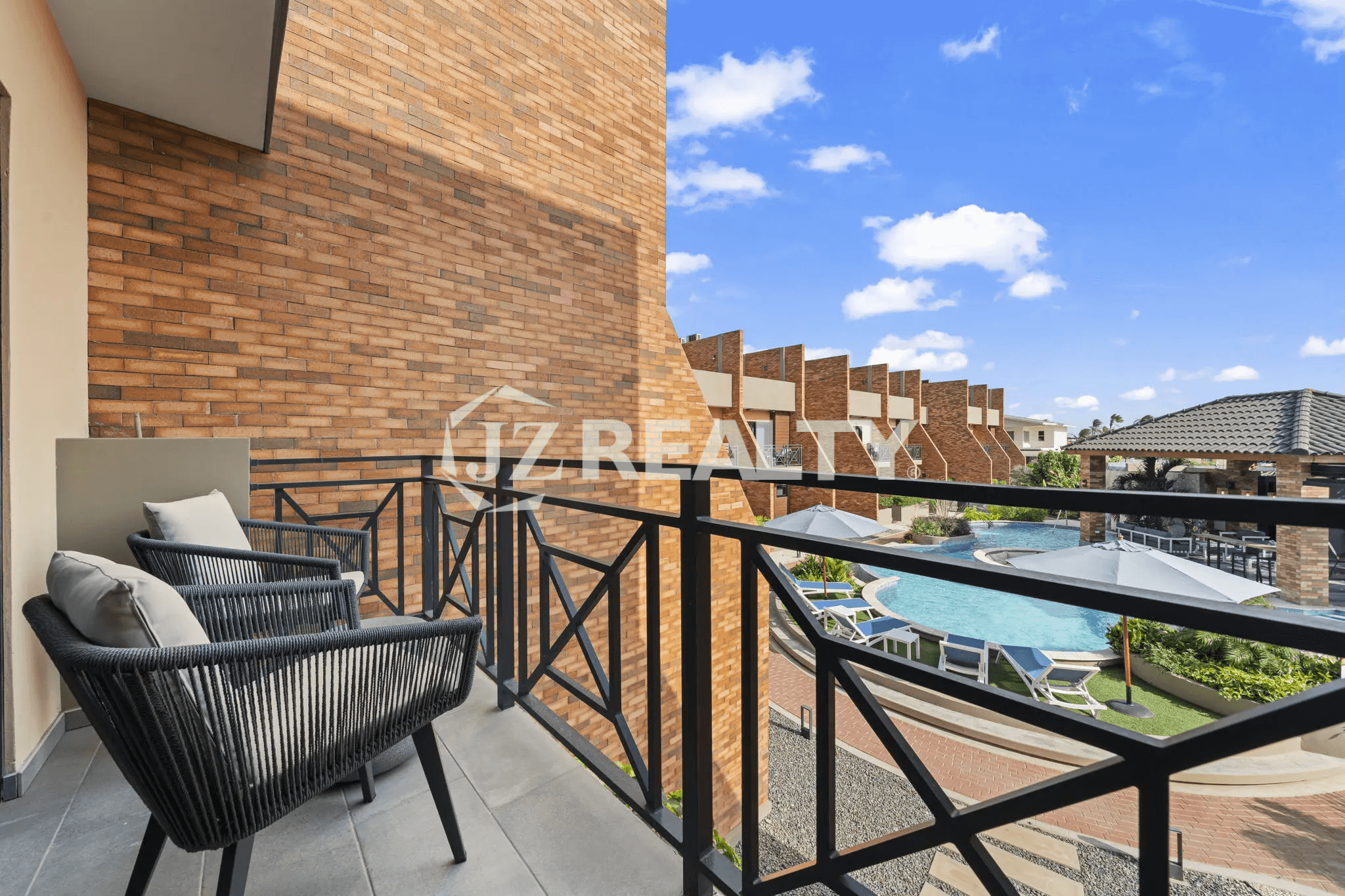 Private outdoor balcony with black metal railings, two modern rope chairs, and a view of the shared pool area and surrounding brick buildings.