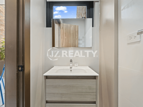 Close-up of a small bathroom sink area with a gray vanity, rectangular mirror, white sink, and white wall tiles.