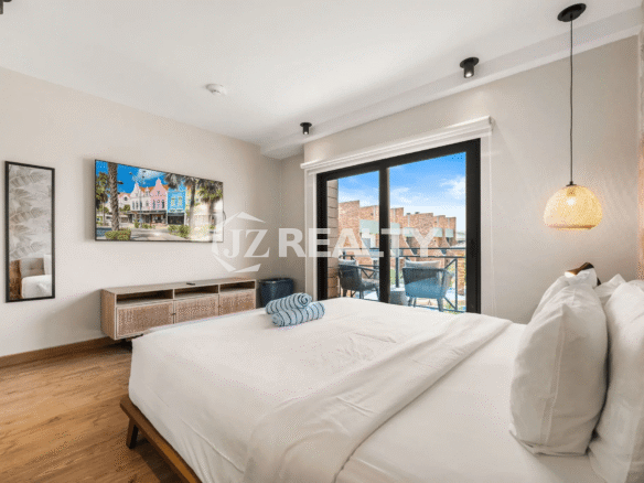 Bright primary bedroom in Porta al Sole Unit 18 with king-sized bed, wall-mounted TV, wood-look floor, and glass sliding door opening to a private balcony overlooking the resort pool area.