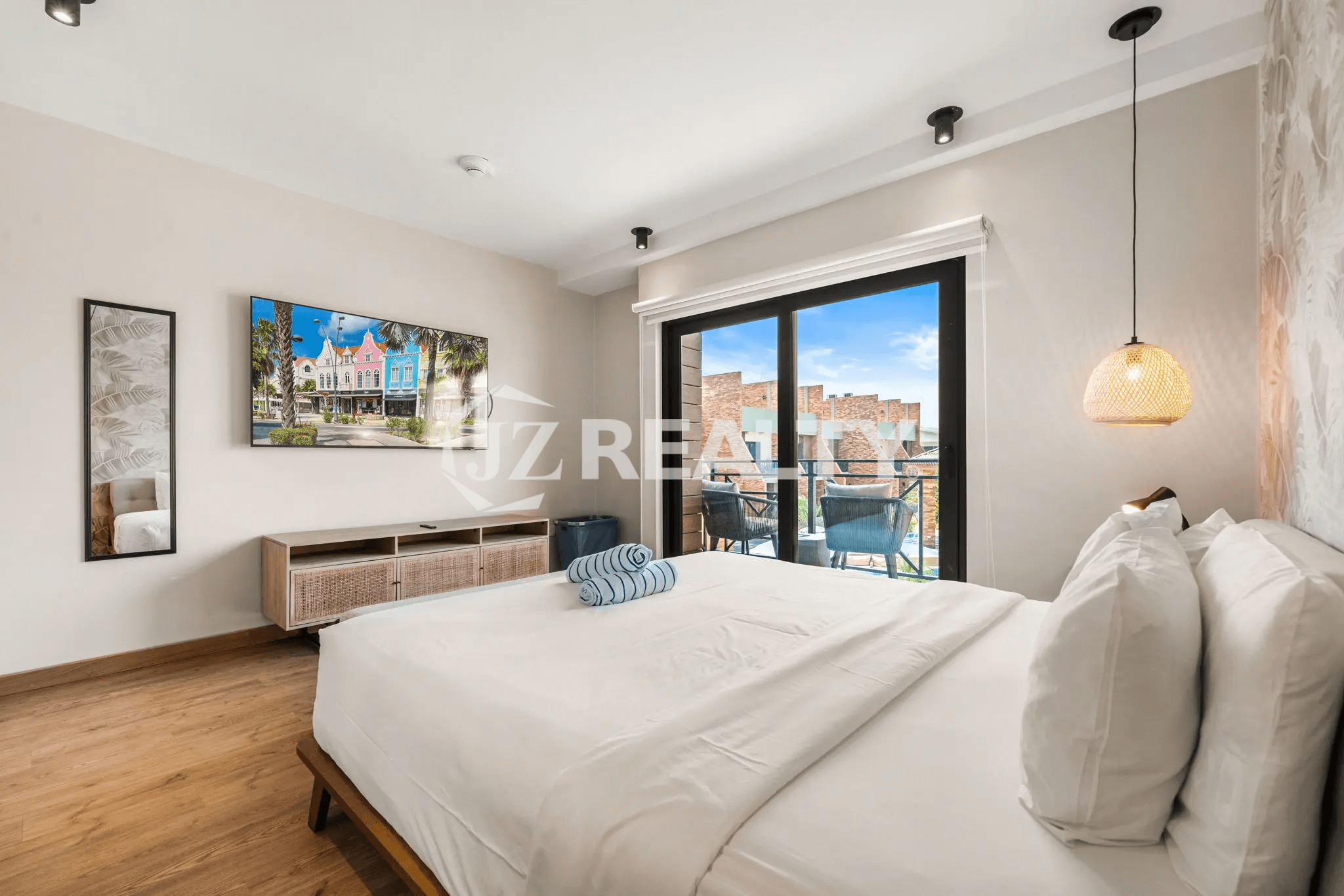 Bright primary bedroom in Porta al Sole Unit 18 with king-sized bed, wall-mounted TV, wood-look floor, and glass sliding door opening to a private balcony overlooking the resort pool area.
