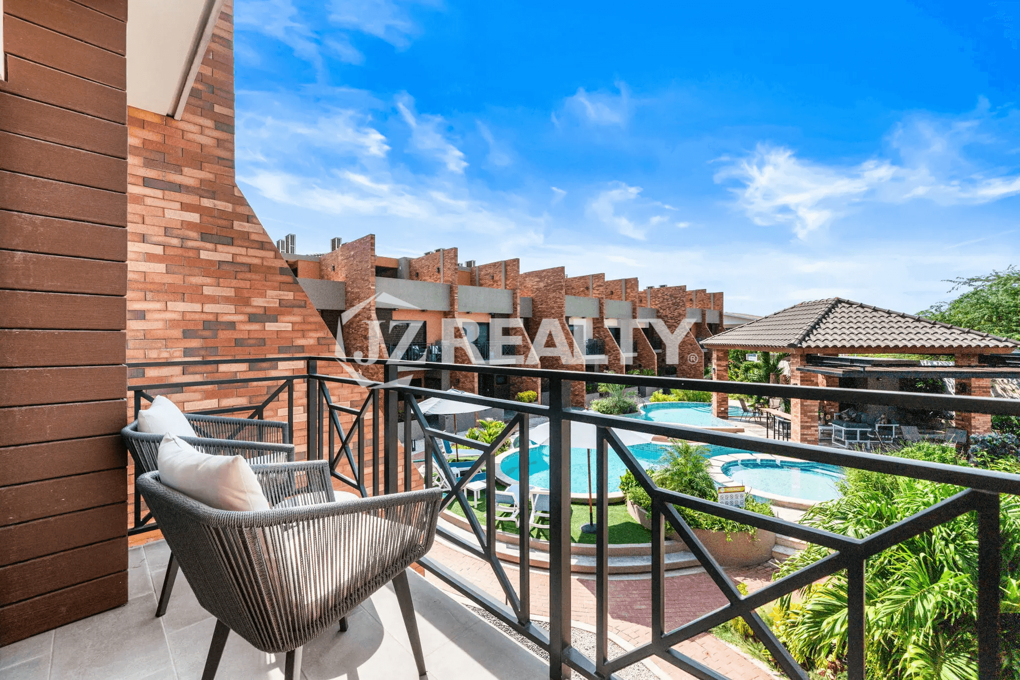 View from the private terrace balcony of Porta al Sole Unit 18, showing a seating area and an expansive view of the community's tropical pool and surrounding brick architecture.