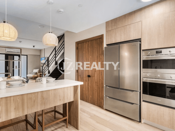 Modern kitchen in Porta al Sole Unit 18 featuring light wood cabinets, stainless steel refrigerator, built-in oven, white countertop island, and rattan pendant lights.