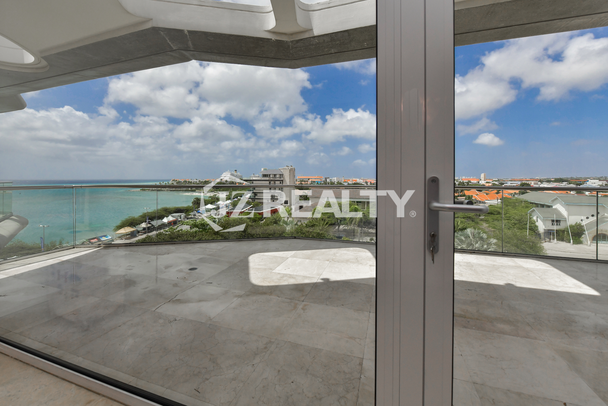 Luxurious balcony at Coral Shell Penthouse B in Aruba, featuring a glass railing, marble flooring, and breathtaking ocean and city views.