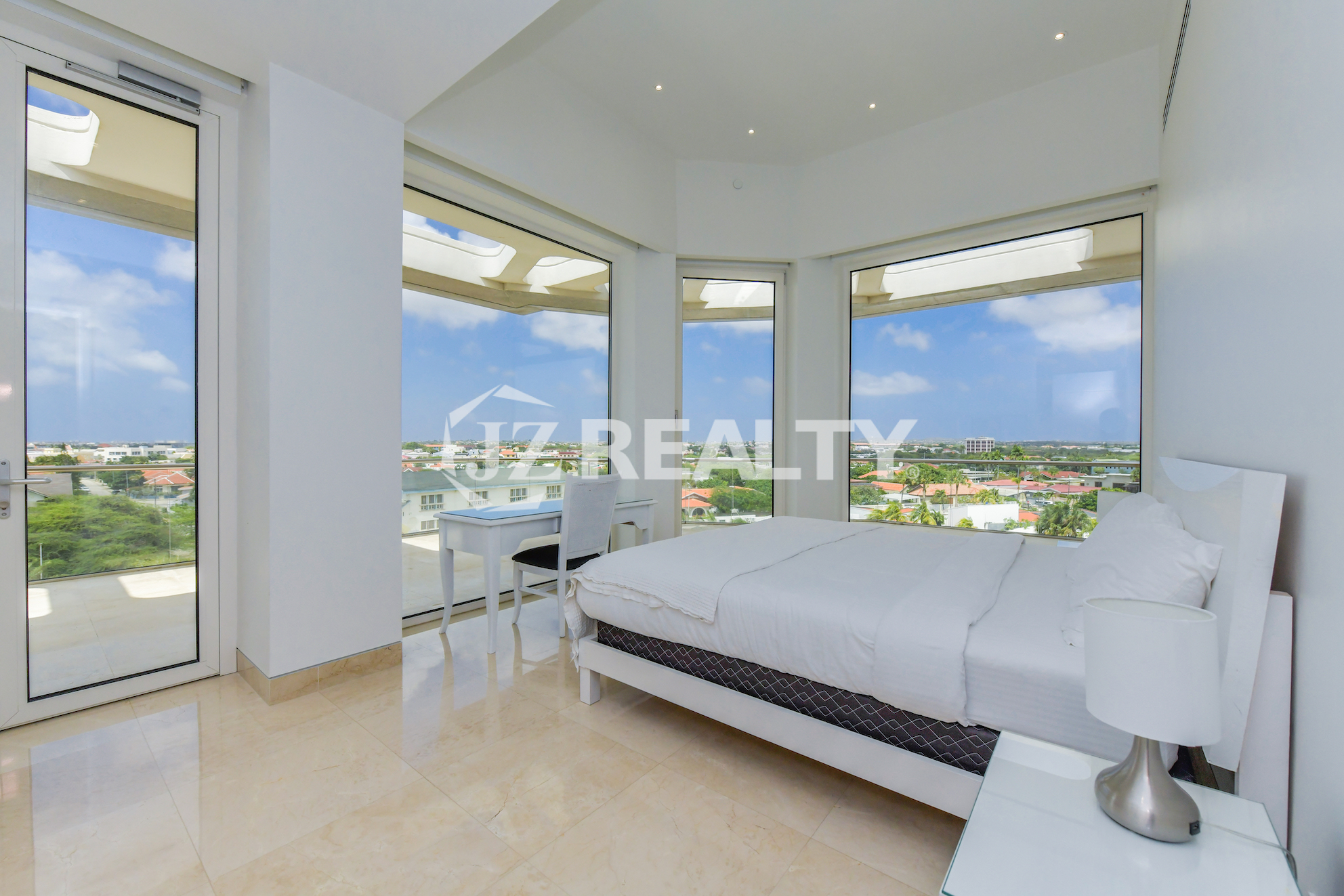 A bright and airy corner bedroom in Coral Shell Penthouse B with floor-to-ceiling windows offering panoramic city and ocean views.