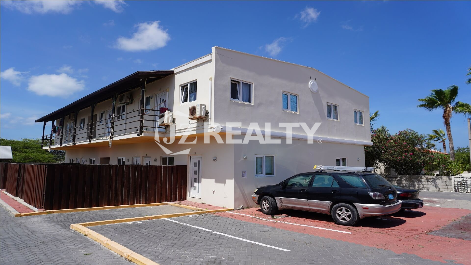 Two-story apartment building with parking bays and upper balconies under blue sky.